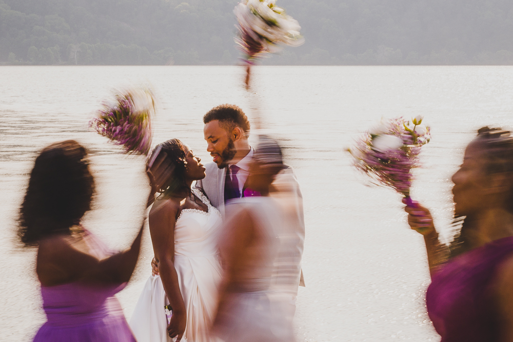 Bride and Groom kissing near a lake in upstate ny in Poughkeepsie with wedding party walking in a blur