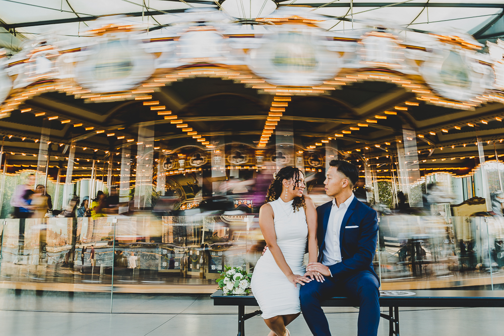 A bride and groom sitting on a bench in front of Jane's carousel in Brooklyn