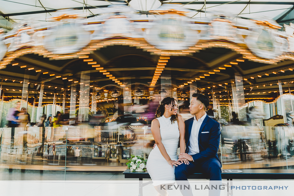 A bride and groom sitting on a bench in front of a carousel.