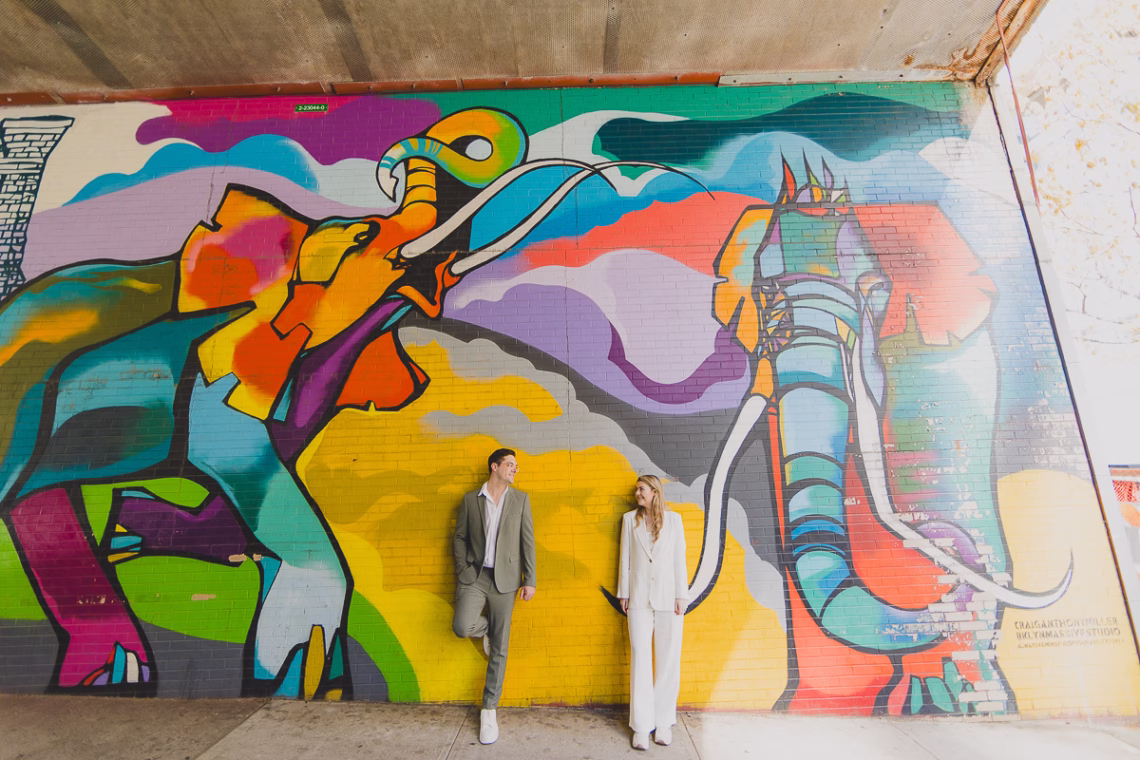 A couple in stylish suits leans against a vibrant, multi-colored brick wall mural featuring two large, stylized elephants. The man wears a sage green suit, and the woman wears a white suit; they are looking toward each other against the bright yellow and purple background.