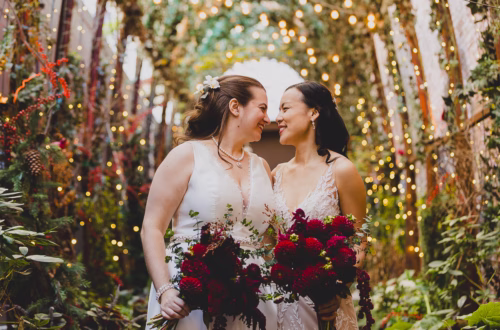 Two brides in white wedding dresses smiling at each other under a greenery arch with twinkling fairy lights, holding bouquets of deep red dahlias.