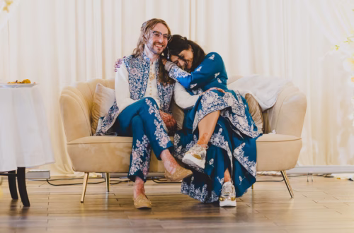Interracial couple in blue outfits sitting on a beige couch embracing and laugh towards each other.