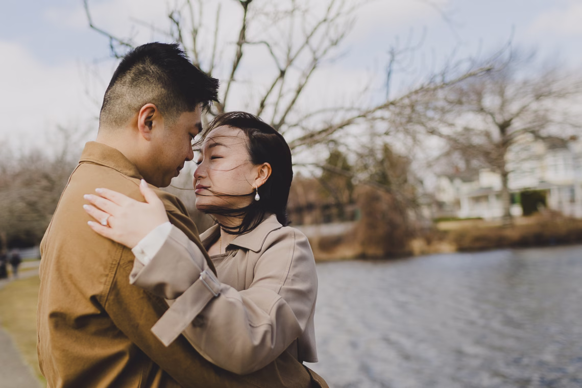 Asian couple in brown outfits almost kissing with their eyes closed near a lake in the background.