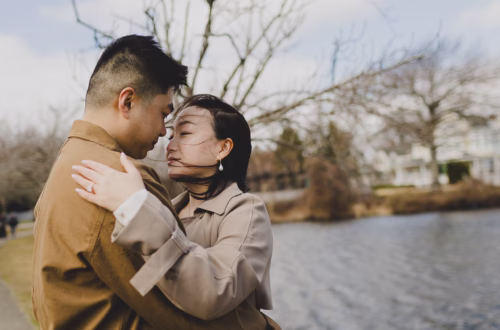 Asian couple in brown outfits almost kissing with their eyes closed near a lake in the background.